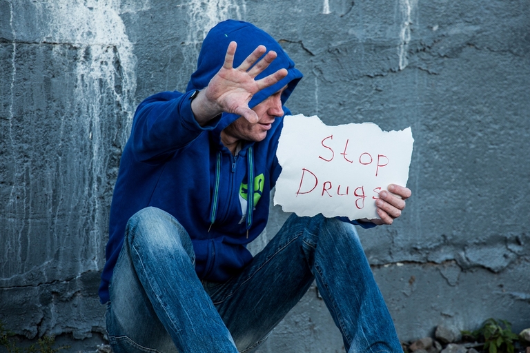 Ein Mann sitzt an einer Mauer und hält ein Schild mit der Aufschrift Stop Drugs
